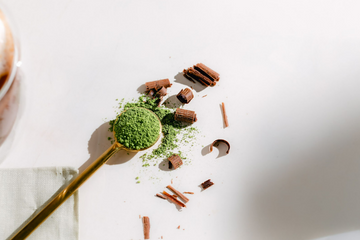 A scoop of matcha powder in a golden teaspoon next to dark chocolate flakes.